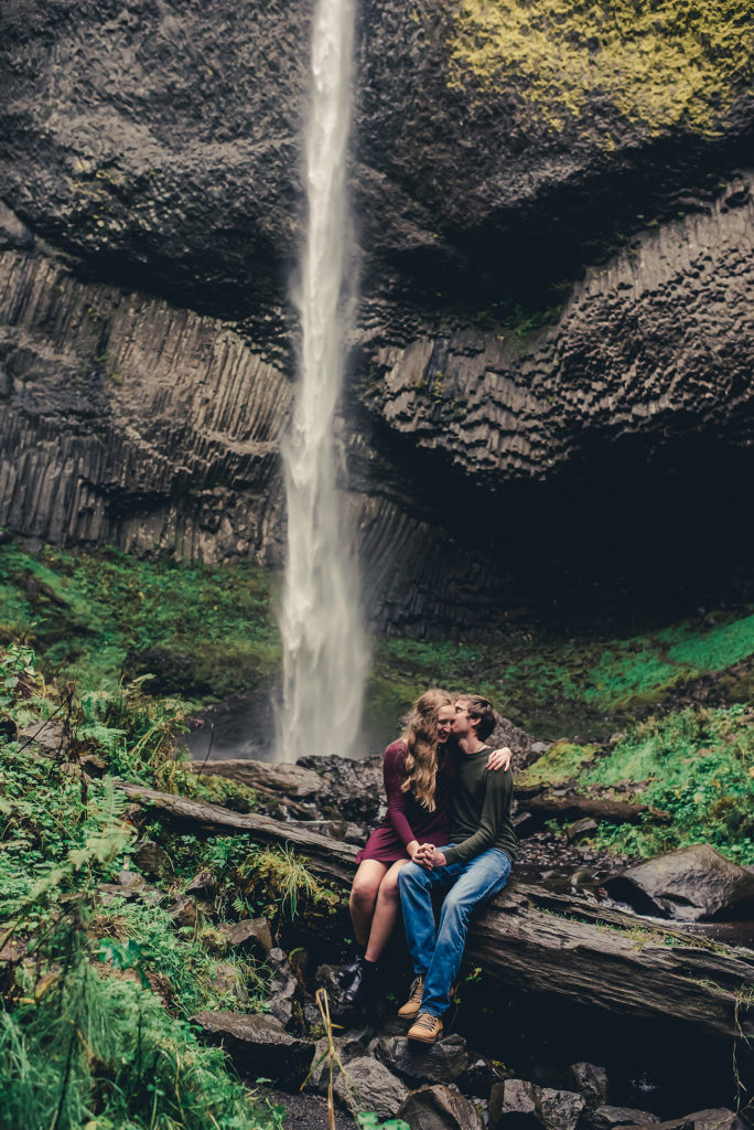 engaged couple posing on log by waterfall
